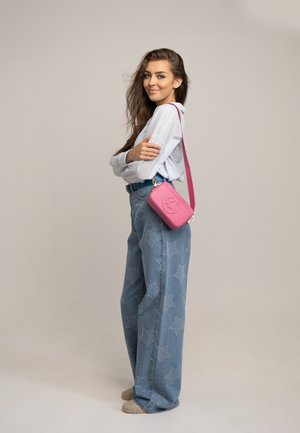 Sac à bandoulière rose avec logo en relief ; chemise bleu clair ; jeans taille haute évasés avec motif étoilé ; chaussures beiges.