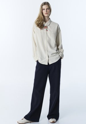 Jeune femme debout, les mains dans les poches, portant une chemise beige à rayures, un pantalon large noir, des baskets blanches et un collier coloré.