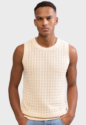 Homme aux cheveux courts et bouclés portant un débardeur en maille beige et un jean bleu clair, les mains dans les poches, fond neutre.