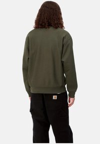 Sudadera de cuello redondo en verde olivo con puños y dobladillo acanalados, llevada sobre pantalones cargo negros con múltiples bolsillos y una etiqueta en la parte posterior.
