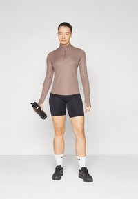 Long-sleeve, collared, brown athletic top paired with black shorts. Model holds a black reusable water bottle, wearing black sneakers and white socks.