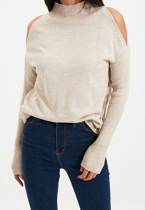 Frau trägt einen beigen Cold-Shoulder-Pullover mit gerippten langen Ärmeln und dunkelblauen Jeans, steht vor weißem Hintergrund.