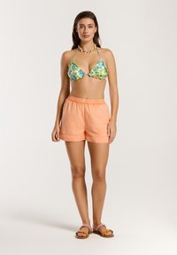 Bikini top s kvetinovým vzorom v zelenej a modrej farbe, skombinovaný s jemnými broskyňovými šortkami a sandálmi s farebnými akcentmi.