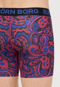 Μάικς σλιπ με μαύρη ζώνη που φέρει το "BJÖRN BORG" σε μπλε. Το ύφασμα παρουσιάζει ένα πολύχρωμο σχέδιο paisley σε κόκκινο, ροζ και μπλε.