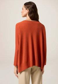 Übergroßer, orangener Pullover aus weichem Material mit gerippten Akzenten. Verfügt über einen weiten Halsausschnitt und einen überschnittenen Schulterbereich. Wird mit hellen Hosen kombiniert.