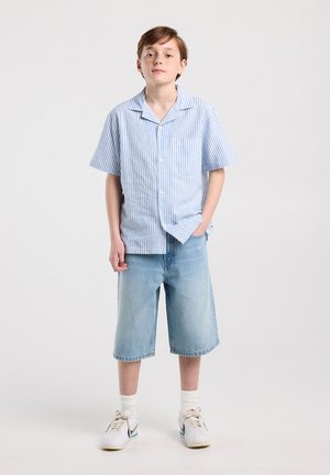 Young person standing with hand in pocket, wearing light blue striped short-sleeve shirt, knee-length denim shorts, white socks, and sneakers.