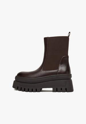 Bota corta de cuero marrón con paneles laterales elásticos, suela plataforma negra gruesa y banda de rodadura resistente. Presenta textura lisa y costuras mínimas.