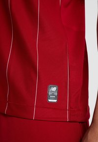 Röd sporttröja med vita vertikala ränder, tillverkad av andningsbart tyg. Har en vävd logotypetikett längst ner till höger.