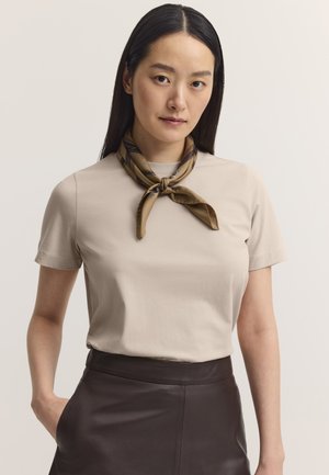 Frau mit langen dunklen Haaren, die ein beiges Kurzarmshirt, einen braunen Lederrock und ein gebundenes braunes gemustertes Halstuch trägt, steht mit einer Hand in der Tasche.