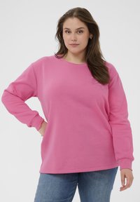 Femme debout avec une main sur la hanche, portant un pull à manches longues rose et un jean bleu, sur fond blanc uni.