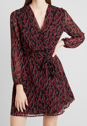 Woman wearing a long-sleeve burgundy and black animal print wrap dress with a waist tie and V-neckline, standing against a plain background.