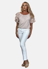 Jasnoróżowa bluzka z falbankami przy rękawach, włożona w białe dopasowane spodnie. Modelka nosi beżowe sandały na koturnie. Jednolity kolor i gładka tekstura.