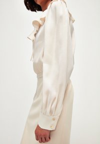 Blusa a maniche lunghe in morbido tessuto beige, con colletto volante, maniche plissettate e polsini con bottoni dorati. Finitura texturizzata sul materiale.