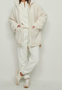 Chaqueta de gran tamaño de crema peluda con un cuello alto, acompañada de una camiseta blanca con cremallera y pantalones de chándal a juego. Zapatillas ligeras con detalles en marrón.