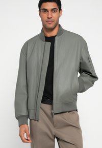 Ανδρικό μπουφάν bomber από γκρίζο δερμάτινο με υφή, με φερμουάρ μπροστά, ριμπ μανσέτες και πλαϊνή τσέπη. Συνδυάζεται με μαύρο t-shirt και μπεζ παντελόνι.