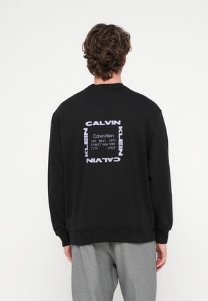 Mann trägt schwarzen Pullover mit Calvin Klein-Logo und New Yorker Adresse auf dem Rücken, kombiniert mit grauer Hose.