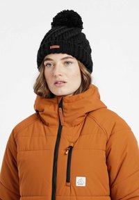 Sort strikket hue med en pompon og et læbelogo, båret med en orange puffjakke med en lynlåslomme foran og hætte.