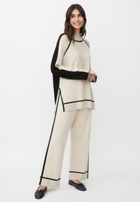 Completo beige a due pezzi composto da una maglia a maniche lunghe con dettagli neri e pantaloni a vestibilità morbida con spacchi laterali e cuciture a contrasto.