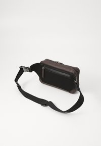 Bolso de hombro negro y marrón hecho de tela texturizada, con cierre de cremallera, correa ajustable y un pequeño bolsillo frontal.