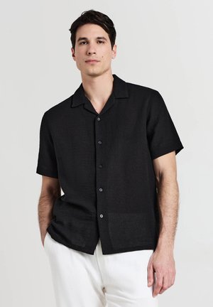 Mann in einem schwarzen kurzärmligen Hemd und weißen Hosen, steht mit einer Hand in der Tasche vor einem schlichten Hintergrund.
