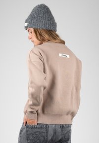 Femme portant un pull beige avec une étiquette dans le dos et un bonnet tricoté gris, regardant vers le bas, avec une main dans la poche d'un jean gris.