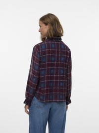 Vrouw zijwaarts kijkend, gekleed in een bordeauxrode en blauwe geruite blouse met lange mouwen en ruches aan de manchetten, gecombineerd met een blauwe spijkerbroek tegen een effen achtergrond.