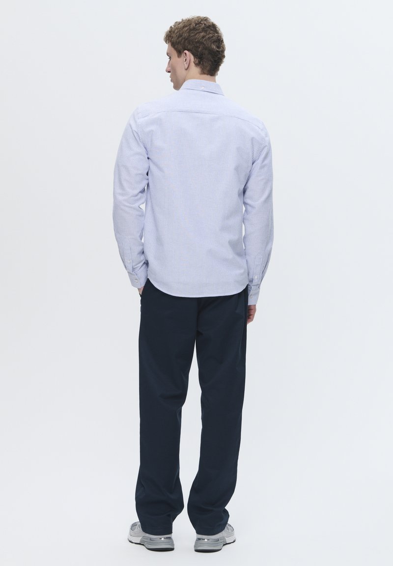 Camisa de rayas azul y blanca, de manga larga con puños abotonados, combinada con pantalones de un oscuro azul marino. El conjunto se completa con zapatos grises.