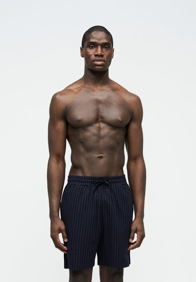 Hombre musculoso de pie, mirando al frente, vestido con pantalones cortos de baño negros con estampado, sobre un fondo claro y liso.