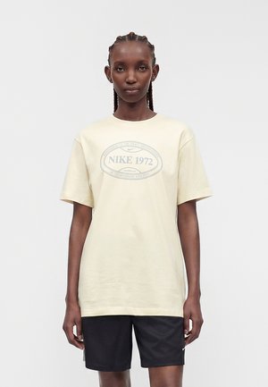 Kobieta z plecionymi włosami, ubrana w kremowy t-shirt Nike 1972 i czarne spodenki Nike, stoi na tle jednolitego jasnego tła.