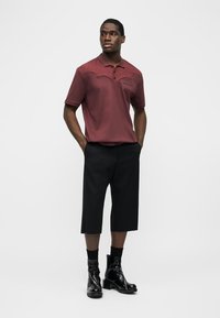 Mann trägt ein kastanienbraunes Poloshirt, schwarze, weit geschnittene, cropped Hosen, schwarze Socken und glänzende schwarze Stiefeletten und steht mit den Händen in den Taschen.