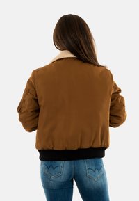 Giacca bomber marrone con colletto in finta pelliccia color crema, polsini e orlo a coste neri, e finitura liscia opaca. Presenta tasche laterali.