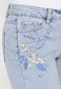 Pantalones vaqueros de mezclilla azul claro con patrones florales en azul, beige y blanco. Presentan detalles metálicos en los bolsillos y una tela suave y texturizada.