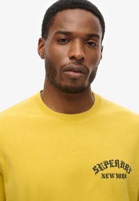 Yellow cotton t-shirt with crew neck, featuring black "Superdry New York" logo on chest. Smooth texture and relaxed fit.