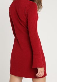 Röd, långärmad miniklänning tillverkad av ett texturerat material. Har utsvängda manschetter och en figurnära silhuett som framhäver en ren, enkel design.