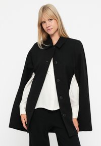 Cappotto in lana nera con colletto tondo, chiusura frontale a bottoni e vestibilità ampia, indossato sopra una camicia bianca e pantaloni neri.