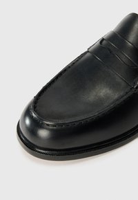 Zwarte leren instappers met een gladde oppervlakte, gedetailleerde stiksels en een ronde punt. Instapmodel met een decoratieve band over de voorkant.