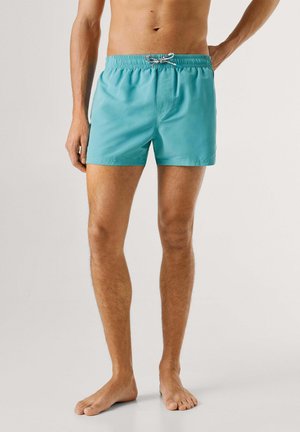 Hombre con pantalones cortos de baño turquesa con cordón blanco y negro, de pie descalzo contra un fondo claro y liso.