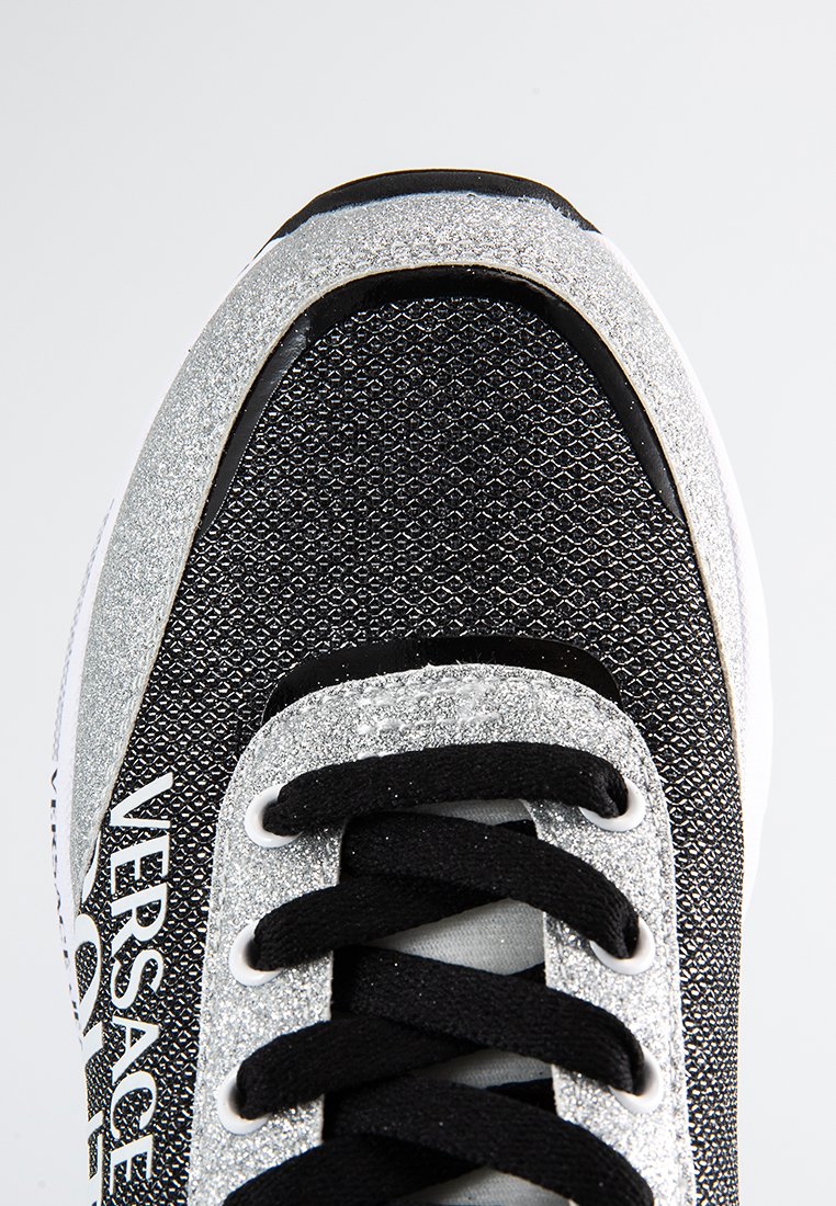 Zapatillas brillantes negras y plateadas con una parte superior de malla texturizada, cordones negros, acentos blancos y detalle del logo en el lado.