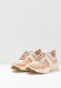 Beige athletic shoes with suede and textile upper, white accents, round toe, and textured rubber sole. Features lace-up closure and pull tab.