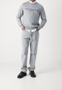 Sudadera gris con logo bordado, camisa blanca con cuello debajo, vaqueros gris claro y zapatos negros. Conjunto casual y de ajuste relajado.