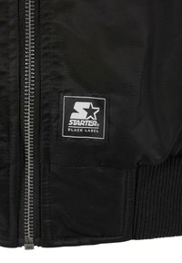 Jachetă bomber neagră cu fermoar metalic, manșete ribbed și o etichetă rectangulară alb-negru cu logo-ul "STARTER BLACK LABEL."