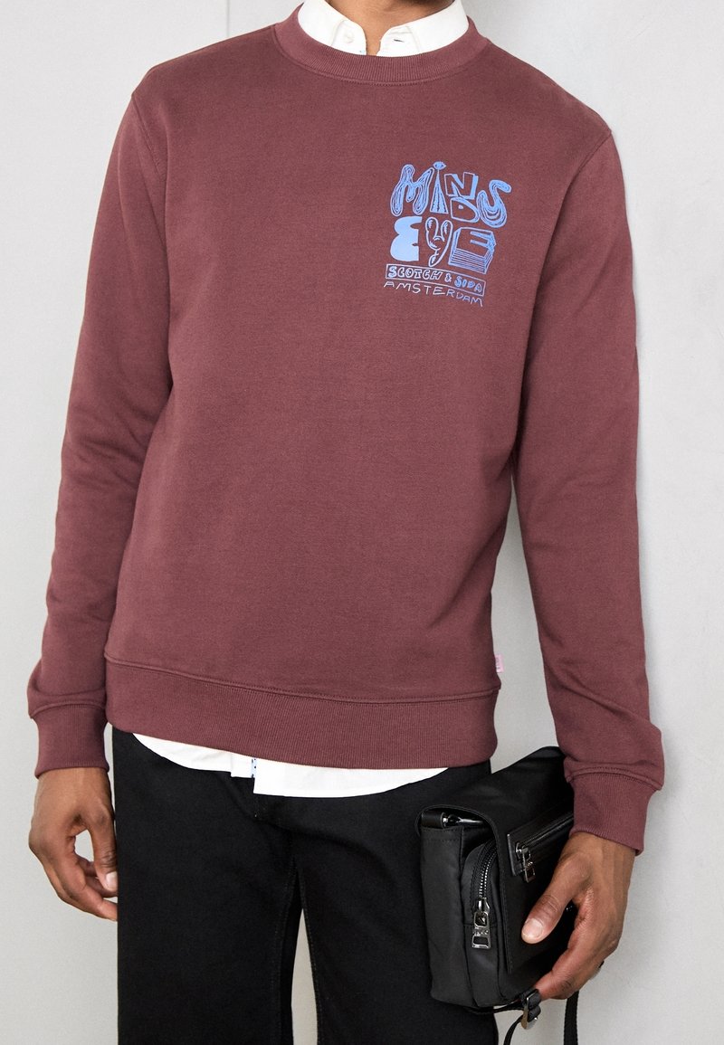 Personne portant un sweat-shirt bordeaux avec un texte graphique bleu, une chemise blanche à col en dessous, un pantalon noir, tenant une pochette noire zippée.