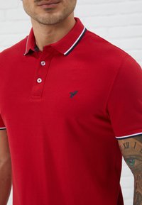 Punainen polo-paita, joka on valmistettu sileästä kankaasta. Siinä on klassinen kaulus, jossa on laivaston sinisiä ja valkoisia raitoja, kaksi nappia ja brodeerattu logo.