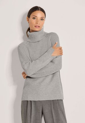 Vrouw in een lichtgrijze geribde coltrui en grijze geplooide broek, staand met gekruiste armen tegen een effen achtergrond.