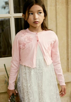 Hellrosa, kurz geschnittene Strickjacke mit floralem Stickmuster und Knopfverschluss, getragen über einem weißen Spitzenkleid mit Perlenakzenten.