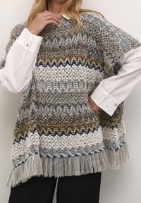 Person som bär en texturerad stickad poncho med zigzag- och popcornstygnsmönster i jordnära färger över en vit skjorta och svarta byxor.