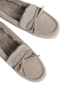 Pantofole mocassino in suede beige con fodera in peluche, punta arrotondata e una decorazione a fiocco sul ponte. Tessuto morbido e dettagli cuciti.