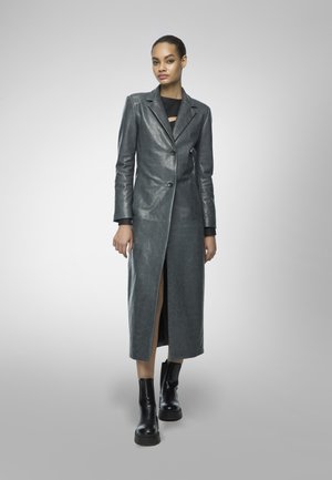 Cappotto lungo in pelle grigia con chiusura a un bottone e un profondo spacco laterale, abbinato a stivali neri alla caviglia e a un top nero aderente.