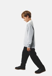 Sudadera gris con un cuello blanco, llevada por un niño en pantalones negros y zapatos negros, mostrado caminando de perfil sobre un fondo blanco.