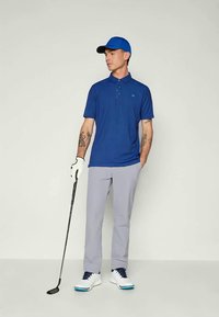 Kék póló, világosszürke nadrág és fehér golfcipő. Golfütőt tart, kék sapkát és fehér kesztyűt visel. Egyszerű dizájn.